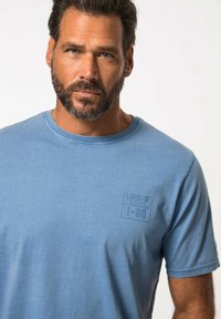 Camiseta de algodón de color azul claro con mangas cortas, que presenta un cuello redondo y un logo estampado en la parte izquierda del pecho. Textura suave.