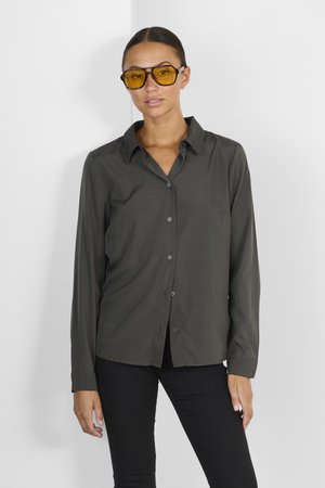 Donkergroene button-up shirt met kraag, lange mouwen en een gladde textuur; gedragen met oversized geelgetinte zonnebril en zwarte broek.
