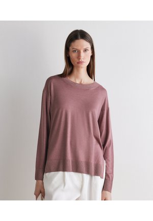 Frau mit glattem braunem Haar, die einen locker sitzenden, mauvefarbenen Pullover mit langen Ärmeln und weiße Hosen trägt, steht vor einer schlichten hellen Wand.