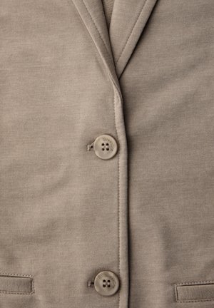 Gros plan sur un blazer en tissu taupe avec deux boutons assortis et des coutures visibles le long des bords du revers et des poches.