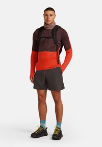 Hombre que lleva una camiseta de manga larga en bloques de color, pantalones cortos oscuros, zapatos de senderismo con cordones amarillos, calcetines azules con patrón, un buff en el cuello y una mochila negra.