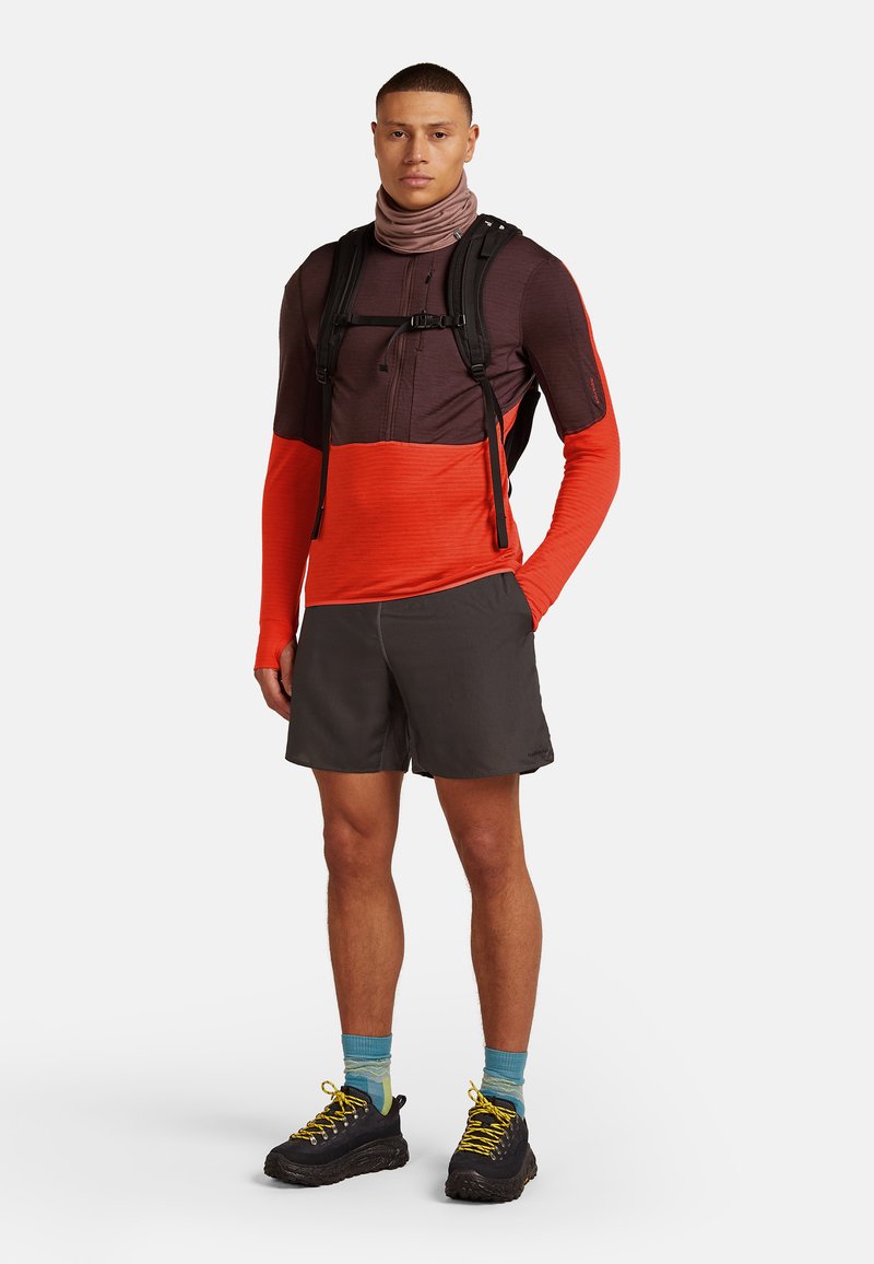 Hombre que lleva una camiseta de manga larga en bloques de color, pantalones cortos oscuros, zapatos de senderismo con cordones amarillos, calcetines azules con patrón, un buff en el cuello y una mochila negra.