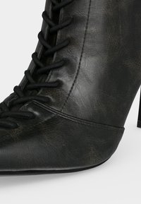 Μαύρο δερμάτινο ankle boot με ψηλό τακούνι, μυτερή μύτη και δέσιμο μπροστά, σε απλό φόντο.