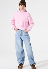 Sudadera corta rosa con texto brillante "FOR", combinada con vaqueros baggy de color azul claro y zapatos marrones con patrón. Fondo sencillo.