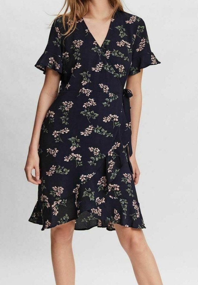 Femme portant une robe portefeuille bleu marine longueur genoux avec imprimé floral rose, manches courtes à volants et un détail lien sur le côté.