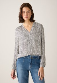 Bluse mit Knopfleiste und V-Ausschnitt, schwarz-weißes Herzmuster und lange Ärmel. Kombiniert mit blauen Jeans aus Denim. Glatte Stoffstruktur.