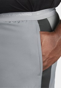 Main dans la poche d'un short de course Nike gris avec taille élastique et panneau latéral noir, porté avec un haut noir.