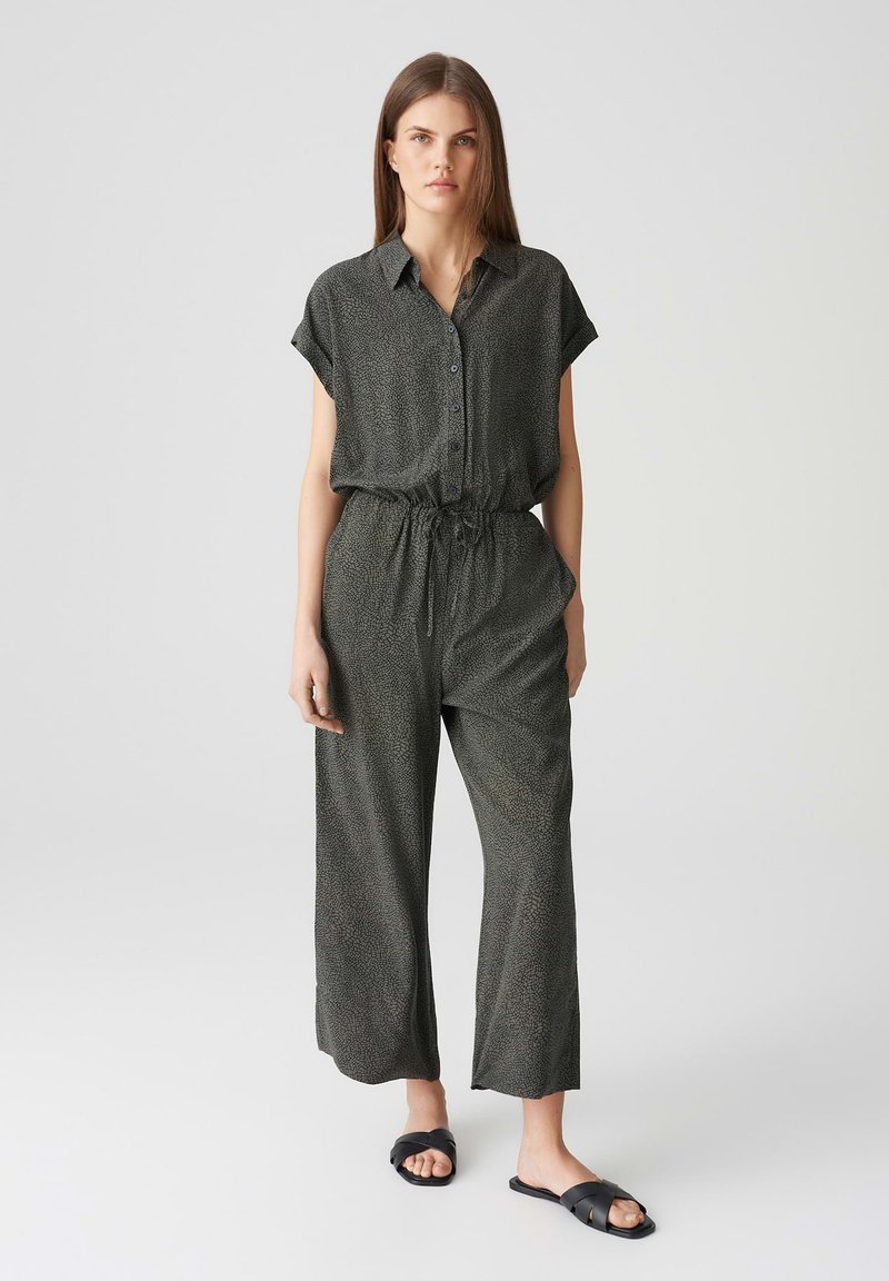 Schwarzer gemusterter Jumpsuit mit kurzen Ärmeln, Knopfleiste, tailliertem Design und weitem Beinausschnitt. Getragen mit schwarzen Rutsch-Sandalen.