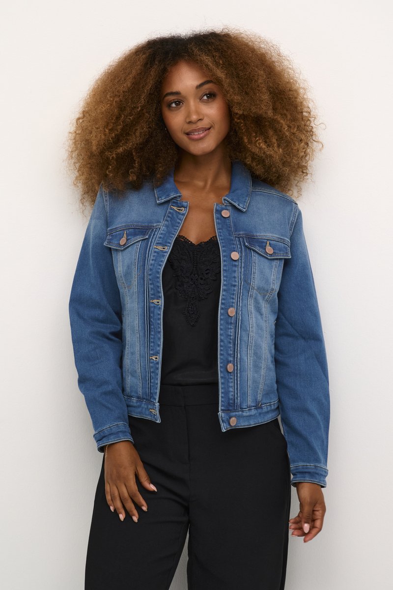 Veste en denim bleu avec manches longues, fermeture à boutons, deux poches poitrine et légère décoloration. Texture douce et coupe décontractée. Portée par-dessus un top noir.