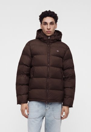 Jeune homme portant une doudoune marron foncé avec capuche, debout devant un fond clair uni, portant un jean bleu clair.