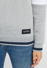 Felpa grigia con accenti bianchi, polsini a costine e fascia blu navy all'orlo. Presenta un logo "oxmo" su un'etichetta nera. Indossata con jeans blu.