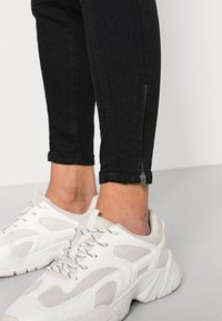 Calças de ganga pretas até ao tornozelo com detalhe em fecho éclair na bainha, combinadas com sapatilhas brancas de malha e sintéticas com detalhes em contraste.