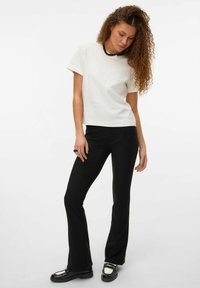 T-shirt en coton blanc à manches courtes associé à un pantalon noir évasé ajusté et des chaussures noires avec un accent argenté. Le mannequin pose légèrement courbé.