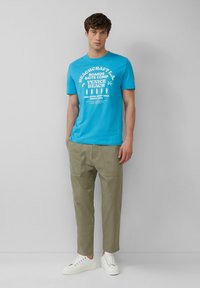 Turqouise katoenen t-shirt met witte grafische tekst, gecombineerd met licht olijfgroene tapered broek en witte sneakers, staande tegen een grijze achtergrond.