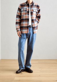 Flanelová košile s mnohobarevným kostkovaným vzorem (modrá, oranžová, černá) a dvěma kapsami na prsou, v kombinaci s modrými džíny a černými botami.
