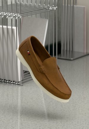 Scarpa marrone in camoscio slip-on con suola bianca che fluttua sopra un pavimento grigio, accanto a un supporto metallico che regge cartelle bianche.