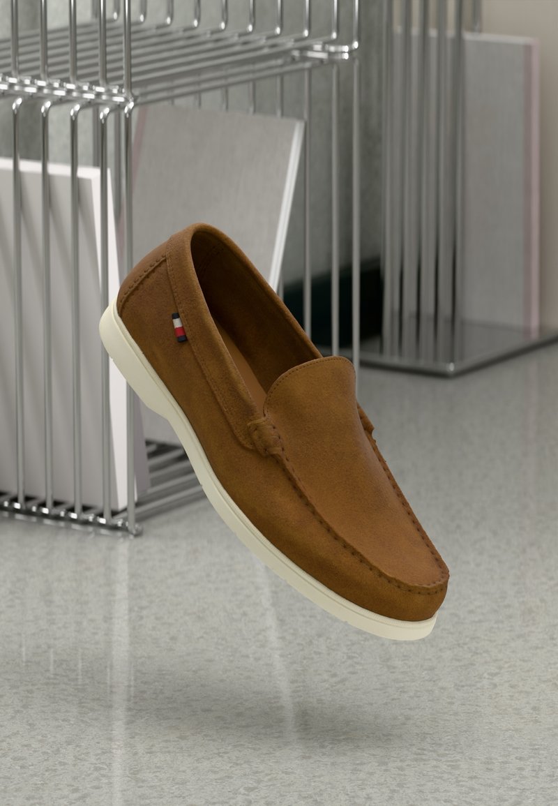 Chaussure à enfiler en daim marron avec semelle blanche flottant au-dessus d'un sol gris, à côté d'un rack métallique avec des dossiers blancs.