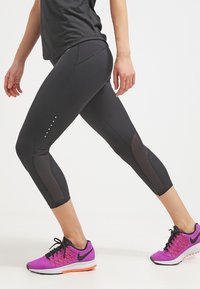 Leggings capri negros con paneles de malla y pequeños puntos de acento en el costado; combinados con zapatillas deportivas rosas y negras. Tela no reflectante.
