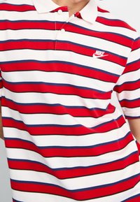 Röd, vit och blå randig polo skjorta med krage, två knappar och en liten Nike-logotyp på vänster bröst. Tyget har en texturerad yta.