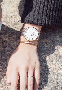 Orologio bracciale in maglia argentata con quadrante bianco, lancette nere sottili e un accento dorato. Tono della pelle liscio e bracciale rigido in oro sottile al polso.