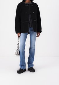 Abrigo de forro polar negro con botones de palanca, suéter gris debajo, jeans acampanados azul claro y botas negras con suela gruesa, sosteniendo una bolsa plateada.