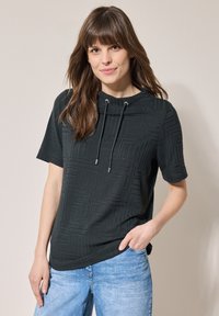 Svart kortärmad topp med ett texturerat geometriskt mönster, med hög hals och justerbar dragsko, kombinerad med ljusblå jeans.