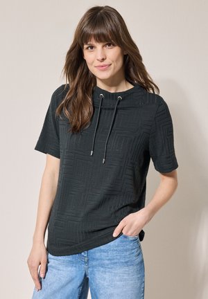 Haut noir à manches courtes avec un motif géométrique texturé, doté d'un col haut et d'un cordon de serrage ajustable, associé à un jean bleu clair.