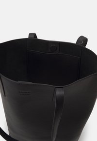 Sac fourre-tout en cuir noir avec une finition texturée, ouverture supérieure et deux poignées latérales. L'intérieur comprend une poche lisse et un marquage discret.