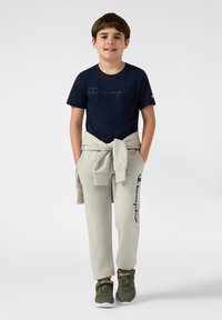 T-shirt Champion a maniche corte blu navy, pantaloni della tuta beige con dettagli del logo e sneakers oliva con cinturino in velcro. Look casual e vestibilità rilassata.