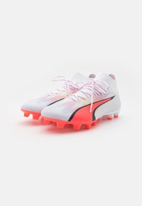 Scarpe da calcio in tessuto bianco con superficie testurizzata, dettagli rosa e neri, e tacchetti arancioni brillanti; presentano lacci a contrasto.