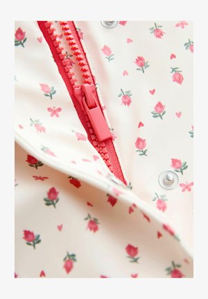 Gros plan sur une fermeture éclair rose et un bouton-pression sur un tissu crème orné de petits motifs de fleurs roses, de cœurs et de papillons.