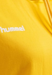 Gul hoodie tillverkad av mjukt tyg, med en vit "hummel"-logotyp, snörning och slät yta.