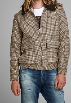 Mann trägt eine beige-braune karierte Jacke mit Reißverschluss und Klappentaschen, kombiniert mit zerrissenen blauen Jeans und einem weißen T-Shirt mit Grafik.