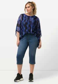 Top transparente azul con estampado, combinado con pantalones cortos de denim de media longitud y zapatillas negras. La parte superior presenta un escote amplio y mangas fluidas.
