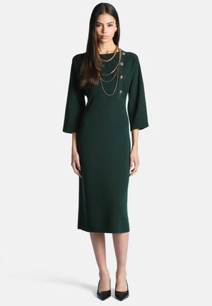 Femme portant une robe midi ajustée vert foncé avec des manches trois-quarts, des boutons dorés sur une épaule, des colliers chaînes dorés superposés et des talons noirs.