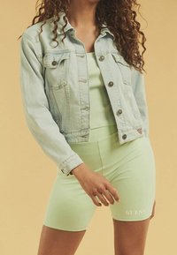 Vaaleansininen farkkutakki napilla, yhdistettynä vaaleanvihreään istuvaan hihattomaan paitoihin ja vastaaviin pyöräilyshortseihin, joissa on valkoinen logo.