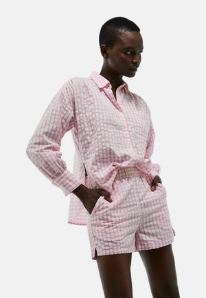 Modèle portant une chemise à manches longues en vichy rose et blanc et un short assorti à taille élastique, les mains dans les poches, regardant sur le côté sur fond blanc.