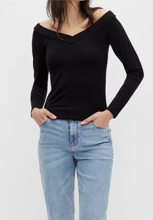 Femme portant un haut noir à manches longues et épaules dénudées avec un jean taille haute bleu clair, les mains dans les poches, sur un fond uni.
