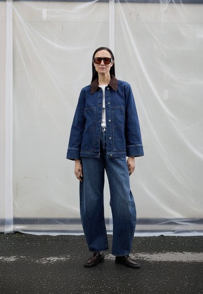 Jeans jakna z rjavim ovratnikom, kombinirana s širokimi jeans hlačami. Model nosi belo majico in velika sončna očala. Teksturirano ozadje.