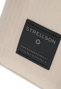 Tessuto beige testurizzato con un'etichetta rettangolare nera recante "STRELLSON" e coordinate. Design semplice con bordo cucito.