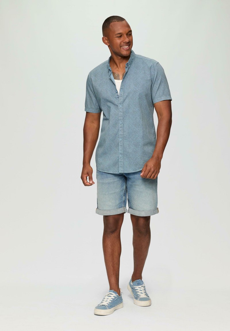 Korte mouwen blauw overhemds met een gestructureerd patroon, gecombineerd met lichte denim shorts en blauwe sneakers. Casual zomeroutfit.