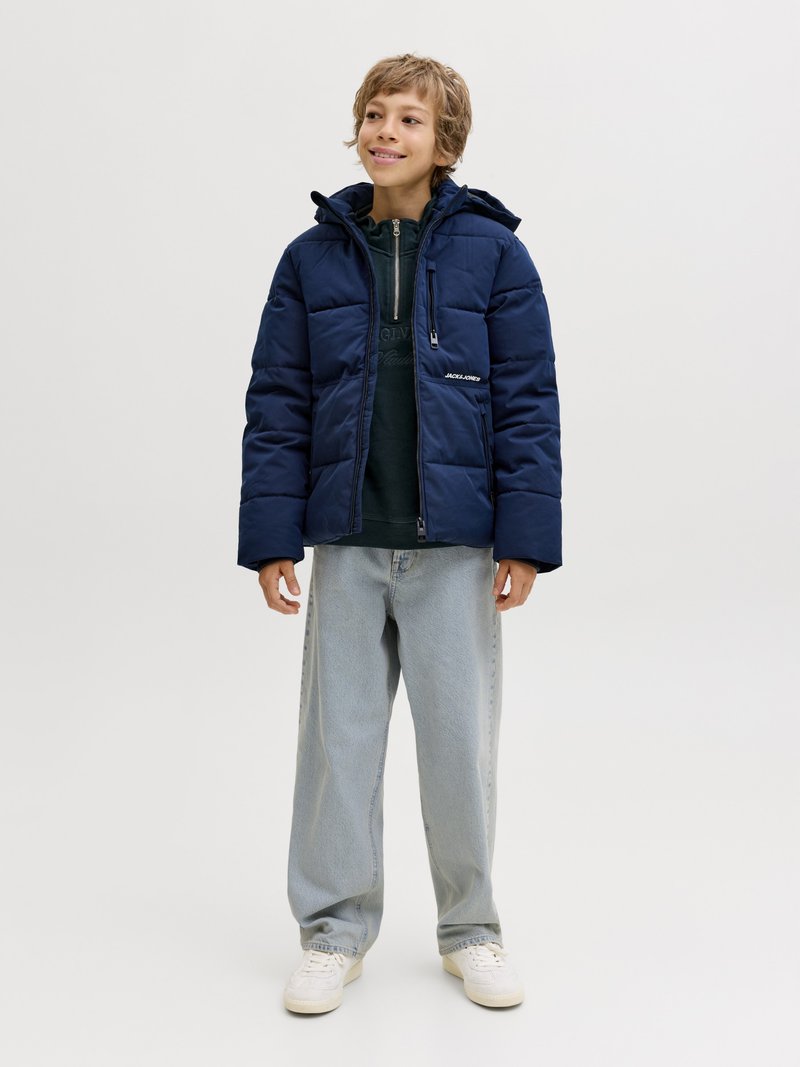 Garçon portant une doudoune bleu marine, un sweat à capuche foncé, un jean large bleu clair et des baskets blanches, debout sur un fond clair uni.