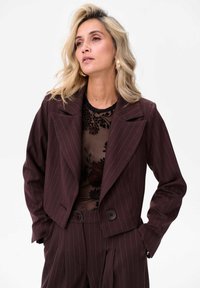 Brązowa marynarka w paski z podwójnym zapięciem i dużym kołnierzem. Nosi się ją na przezroczystą, kwiecistą bluzkę. Marynarka ma ozdobne guziki.