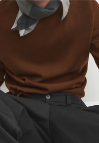 Bruine gebreide trui met een ronde hals, gecombineerd met een grijze sjaal met een patroon. Zwarte broek met een knoop en een strak ontwerp.