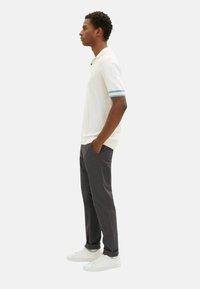 Poloshirt in Weiß mit blau-gelben gestreiften Ärmeln, kombiniert mit geraden dunkelgrauen Hosen und weißen Sneakern, im Profil dargestellt.