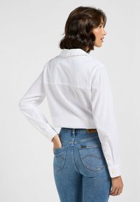 Weißes, kurzes Hemd mit langen Ärmeln und Knopf-Akzenten, kombiniert mit hoch taillierten blauen Jeans, die über Gesäßtaschen und helle Nähte verfügen.