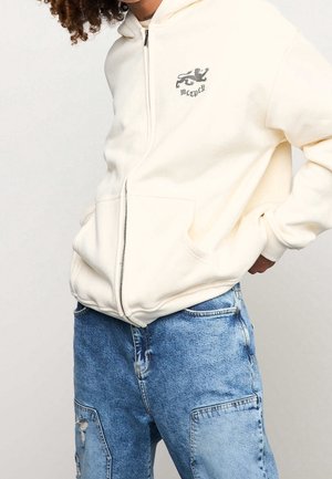 Person wearing cream zip-up hoodie with black lion emblem and text on chest, paired with blue patchwork denim jeans.