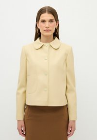 Beige Jacke ohne Kragen mit einer kastenförmigen Silhouette, ausgestattet mit einer Vordertasche und runden Knöpfen. Eine glatte Textur, kombiniert mit einem braunen Rock.