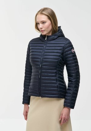 Donna che indossa una giacca imbottita trapuntata blu navy con zip, cappuccio e patch con logo sulla manica, abbinata a una gonna beige.