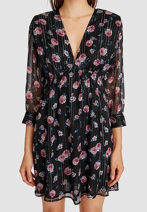 Robe longue noire transparente à manches longues avec motif floral rose et bleu, décolleté en V profond et taille cintrée, portée par une personne debout.
